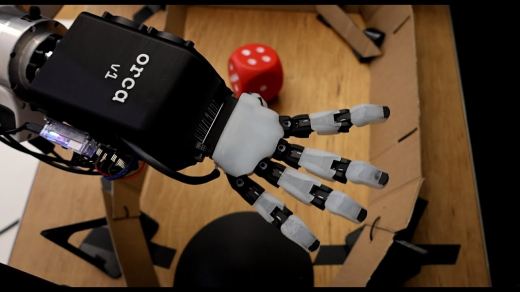 Mano robotica ORCA v1 con dita antropomorfe in posizione aperta, rivolta verso un dado rosso su una superficie in legno