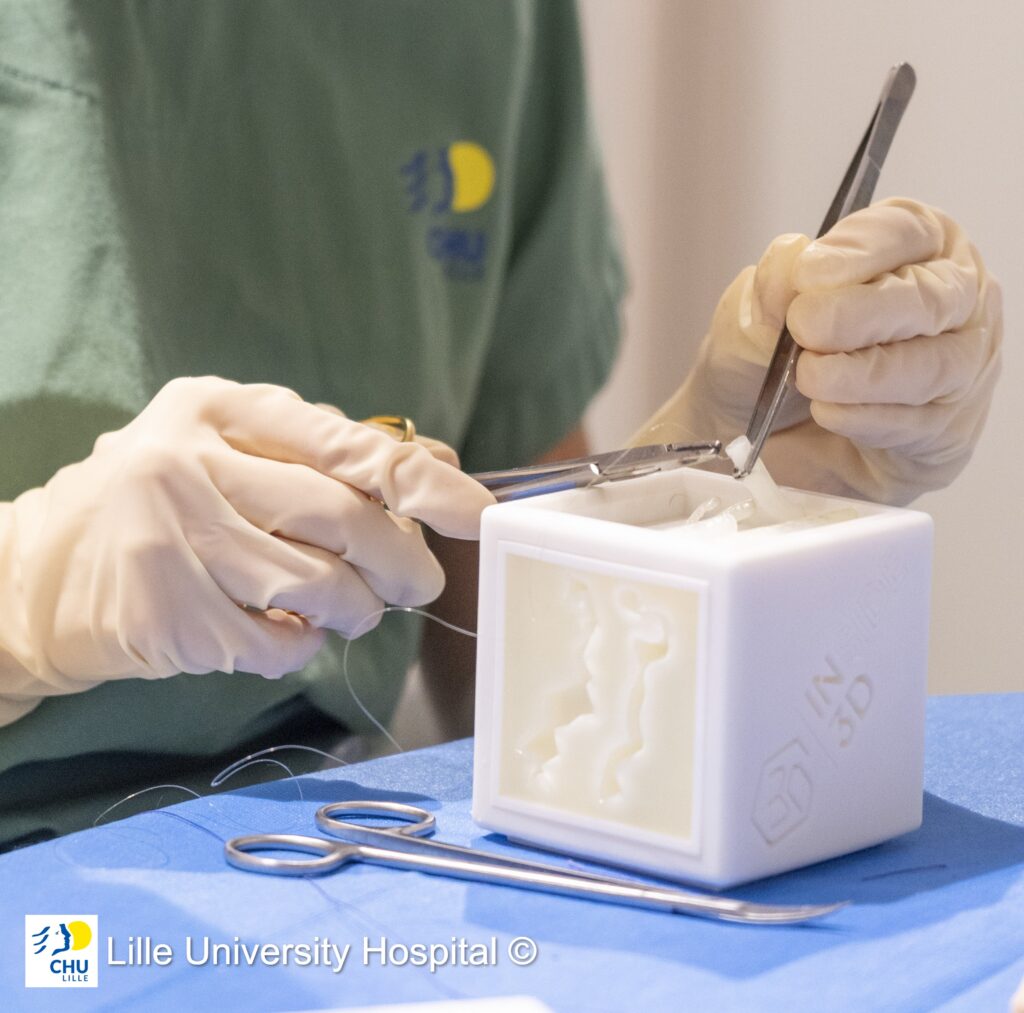 Mani in guanti chirurgici utilizzano pinzette e porta-aghi per esercitarsi nella sutura su un cubo stampato in 3D